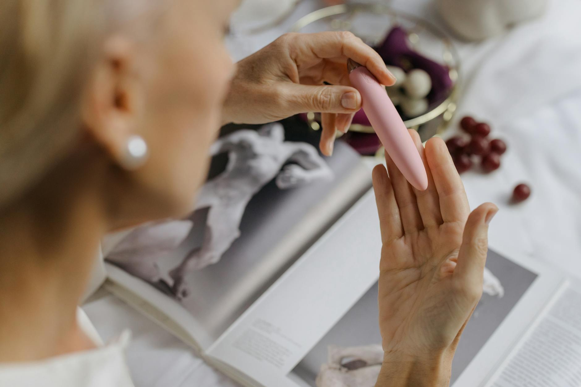Woman holding a pink vibrator while reading, surrounded by art books, representing intimate pleasure and mental presence