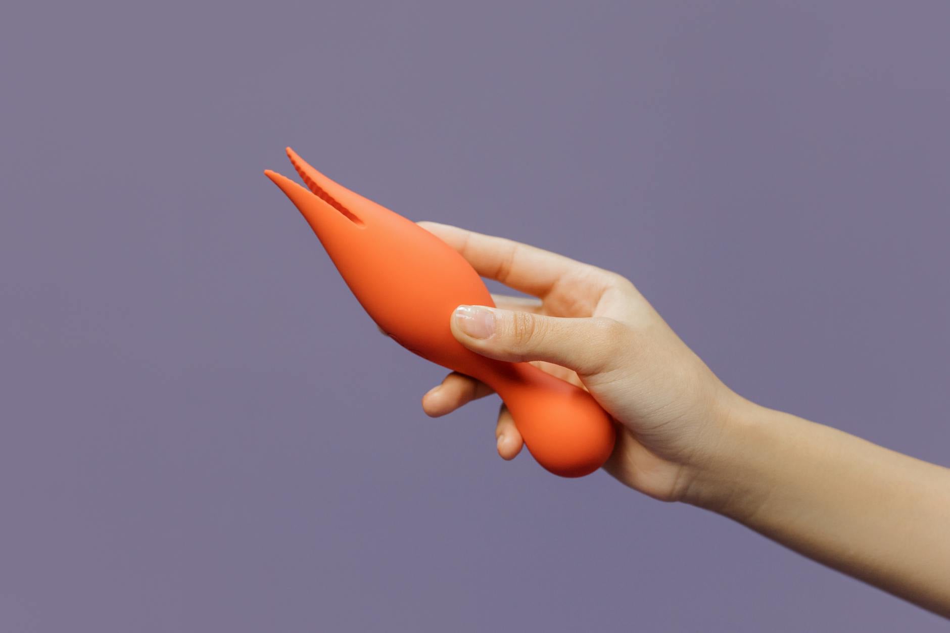 Close-up of a hand holding an orange clitoral vibrator against a minimalist backdrop, representing gentle postpartum pleasure recovery.