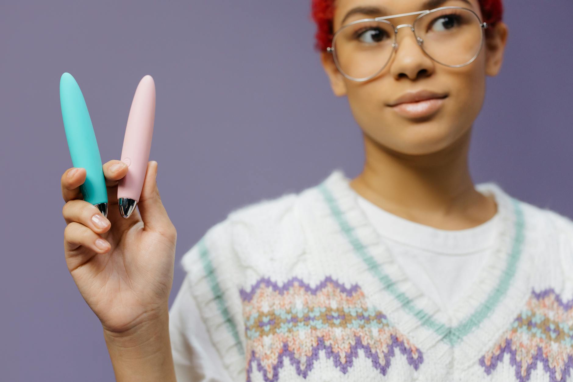 A person with eyeglasses holding colorful clitoral vibrators against a purple background, symbolizing pleasure exploration and choice.