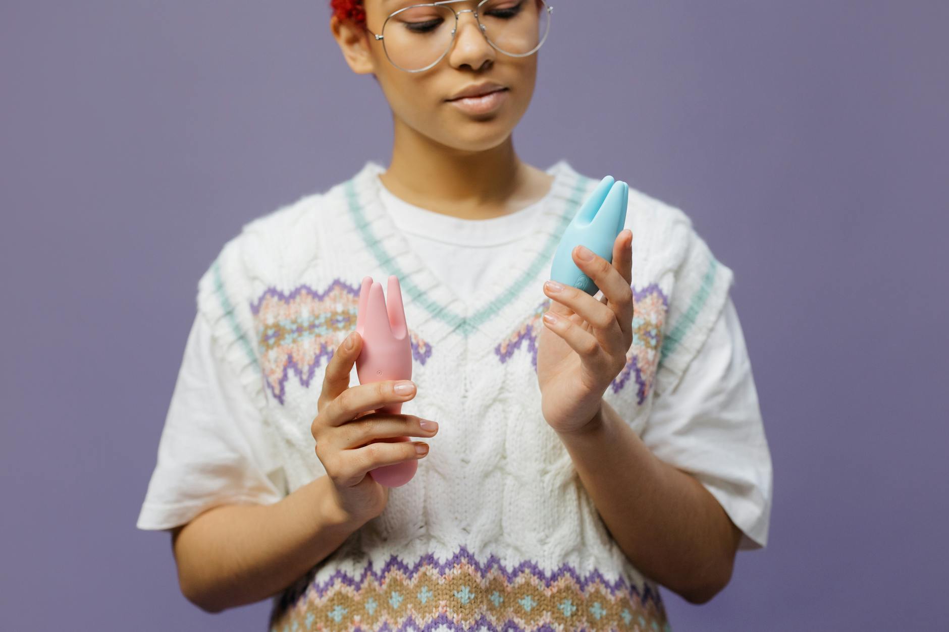 Woman with eyeglasses holding blue and pink clitoral vibrators in a contemplative manner.