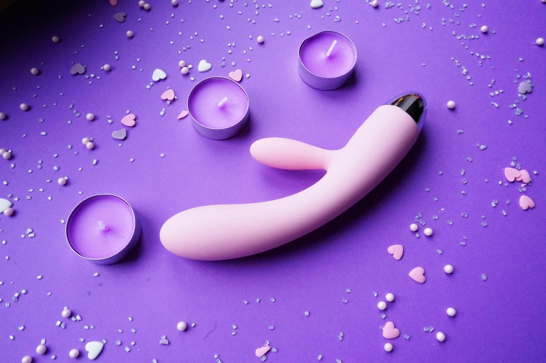 Pink vibrator surrounded by candles and heart confetti on a purple background, symbolizing intimacy and reconnection.